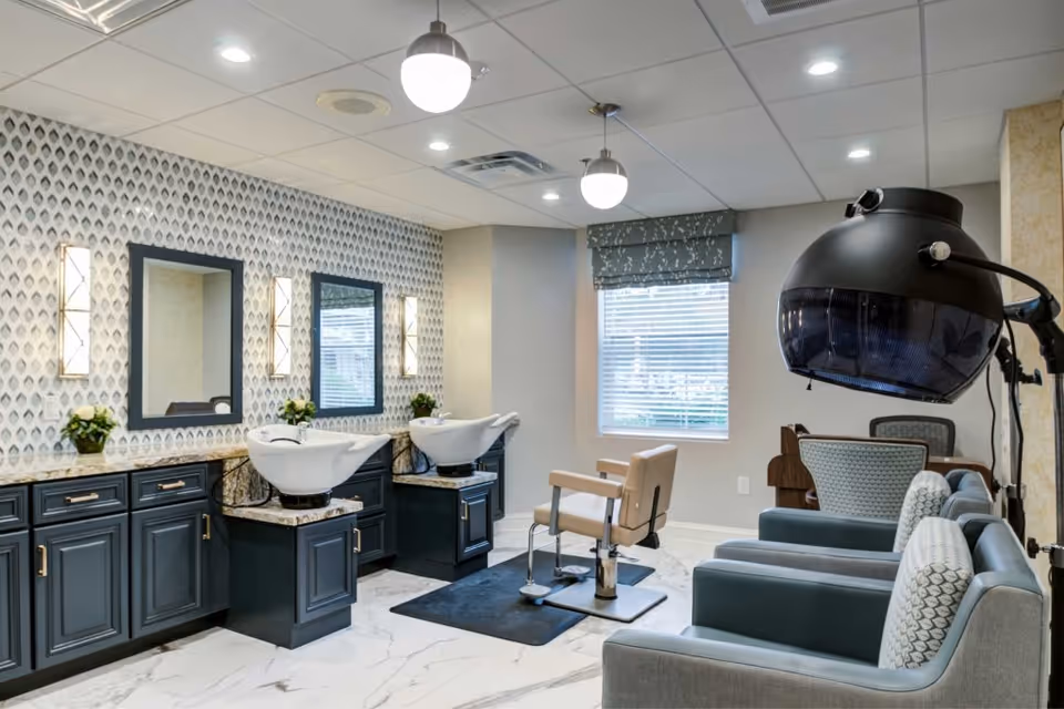 Interior of a salon area in a senior living facility featuring two washbasins with mirrors and wall lights above dark cabinetry, a beige salon chair on a black mat, a large window with a patterned valance, and a black hair dryer hood next to two blue armchairs with patterned cushions.