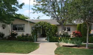 Single-story care home exterior with walkway, trees, and landscaped garden.