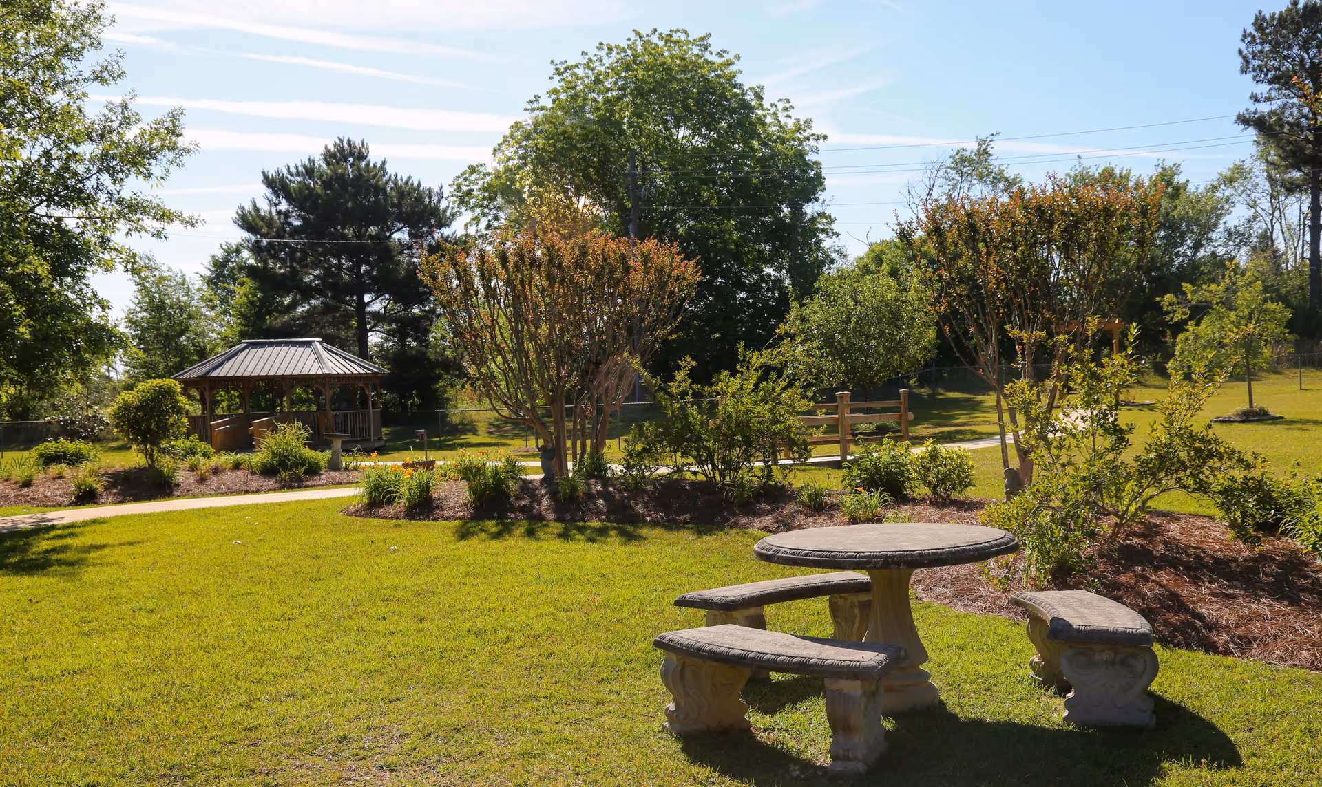 A sunny outdoor garden area with a stone table and three curved stone benches on a grassy lawn. There are various bushes and trees surrounding the area, and a wooden gazebo is visible in the background along a paved pathway.