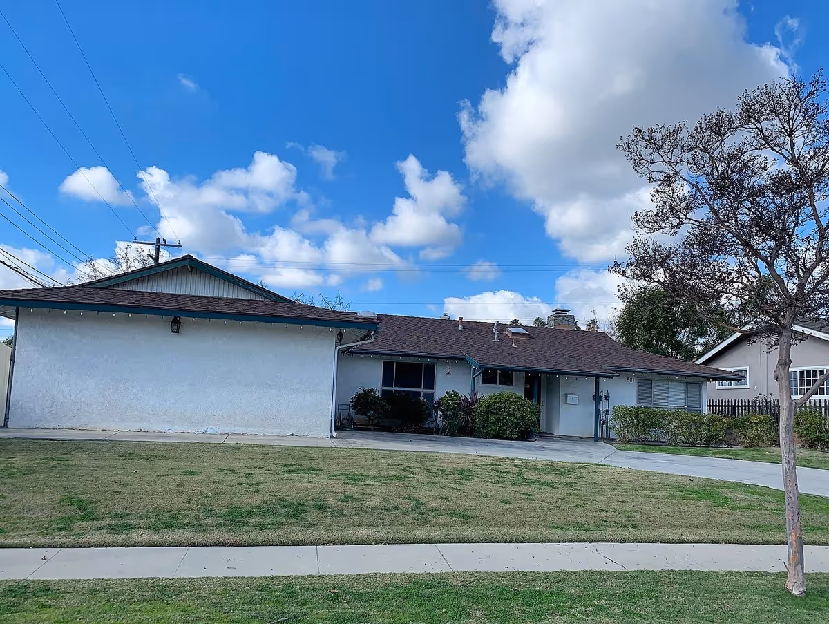 Single-story residential building with a sloped roof, white exterior walls, and a small covered porch area. The front yard has a patchy lawn and a sidewalk, with a leafless tree on the right side. The sky is blue with scattered clouds.