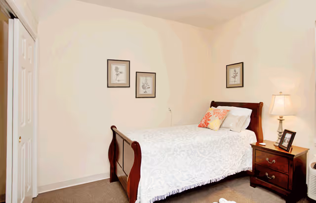 A small bedroom with a wooden sleigh bed covered in white bedding, a nightstand with a lamp and framed pictures on the wall.