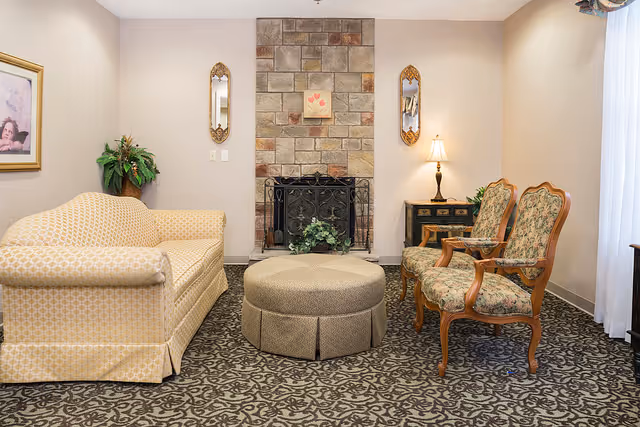 A cozy living room with a patterned carpet, a beige patterned sofa on the left, two floral upholstered wooden armchairs on the right, and a round beige ottoman in the center. The back wall features a stone fireplace with a black metal screen, flanked by two decorative mirrors and a small framed artwork above the fireplace. A table lamp and a small plant sit on a dark wooden side table to the right of the fireplace.