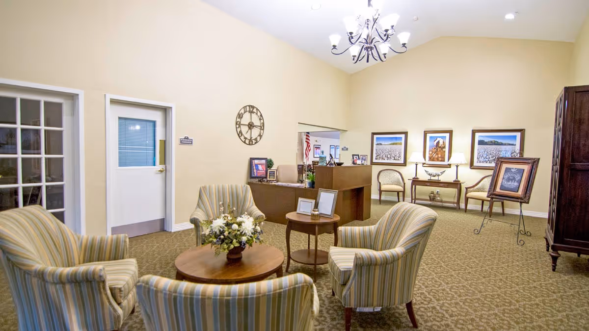 A cozy waiting or reception area with four striped armchairs arranged around a round wooden coffee table with a flower arrangement. The room has beige walls, a carpeted floor, a chandelier hanging from the ceiling, and framed pictures on the walls. There is a reception desk in the background with some chairs and decorative items, and a door with a window on the left side.