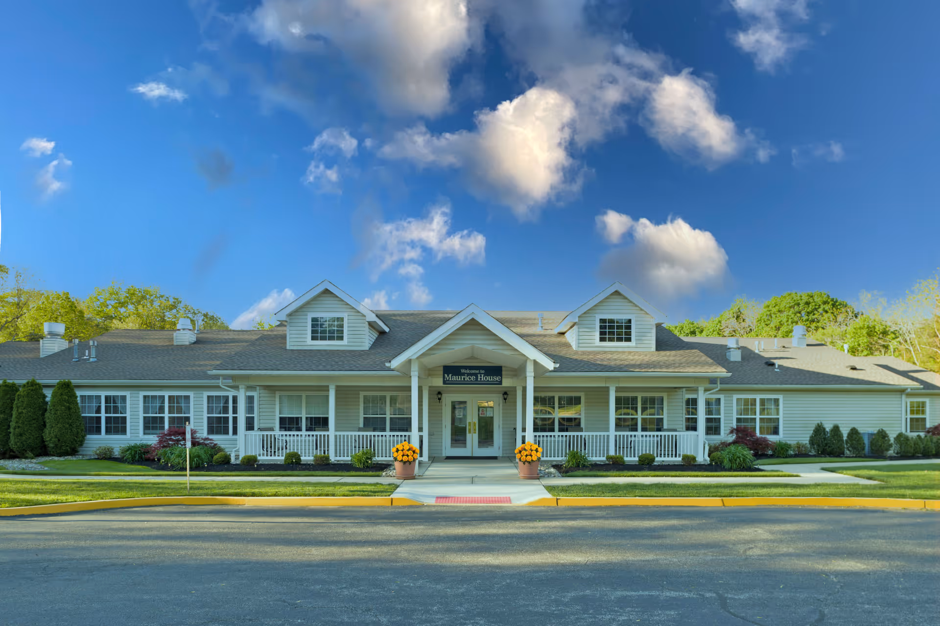 Front exterior view of Maurice House Assisted Living Community building with a welcoming entrance, white railing, potted flowers, and a clear blue sky with some clouds.