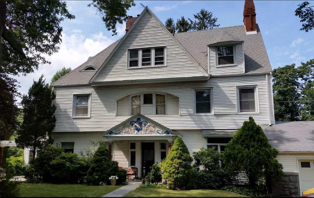 Front exterior view of a large, white, multi-story house with a steep roof, multiple windows, a small porch with decorative trim, surrounded by green trees and shrubs under a partly cloudy sky.