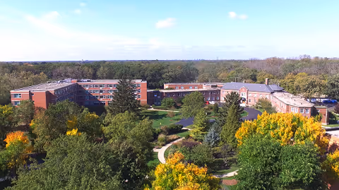 Aerial view of Cantata Adult Life Services a CCRC facility surrounded by lush green trees with some trees showing autumn colors. The large brick building complex is set amidst a landscaped area with pathways and open green spaces under a clear blue sky.