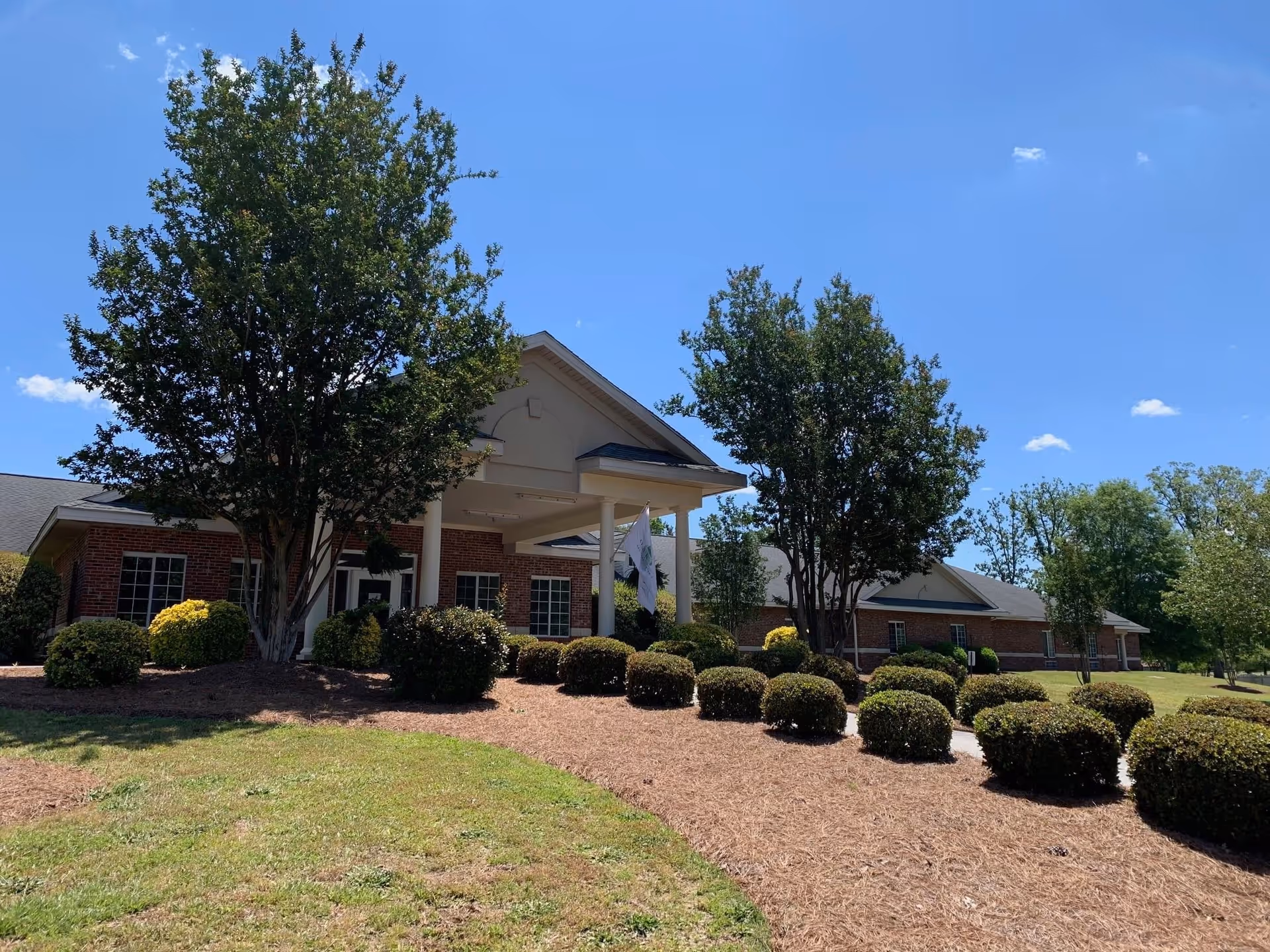 Exterior view of Oak Hill Assisted Living facility showing a brick building with a covered entrance, surrounded by neatly trimmed bushes and trees under a clear blue sky.