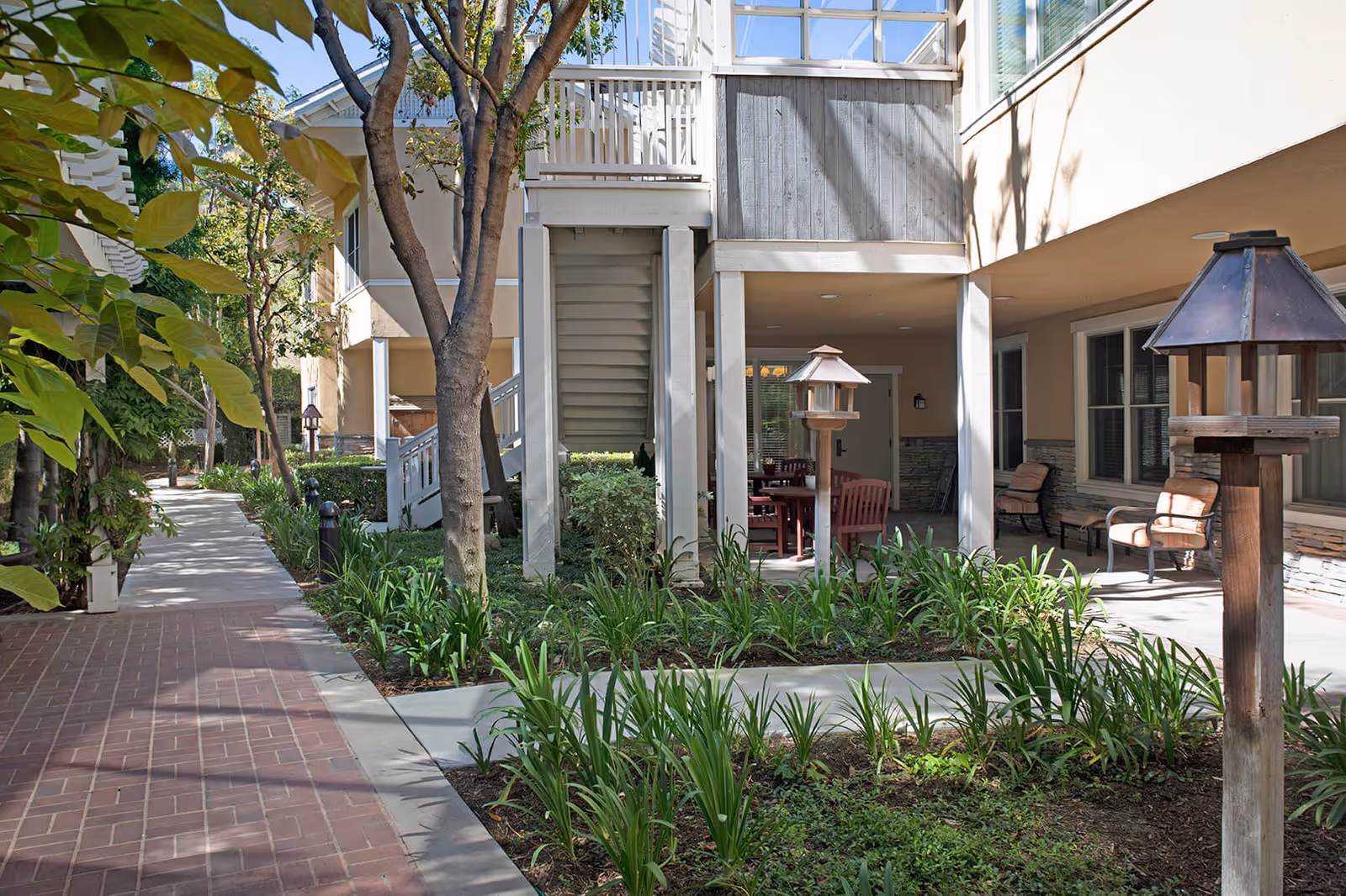 Outdoor view of a senior living facility showing a walkway lined with plants and trees. There are two wooden birdhouse-style lanterns on posts, and a covered patio area with chairs and a table. The building has beige walls and large windows, with stairs leading to an upper level.
