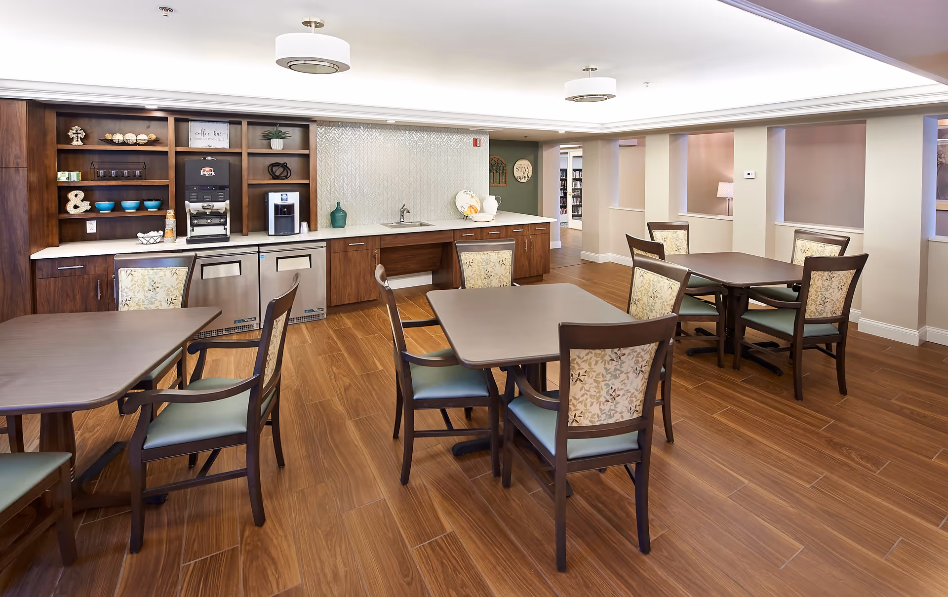 A bright and clean dining area with wooden floors, several square tables each surrounded by four chairs with floral upholstery. The back wall features a countertop with a sink, coffee machines, and shelves holding decorative items and cups. The room has recessed lighting and a modern ceiling design.