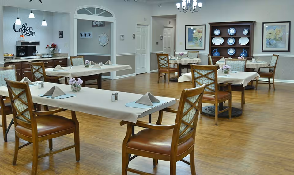 Spacious assisted living dining room with several tables set with napkins, wooden chairs, a coffee bar, and a dish display cabinet.