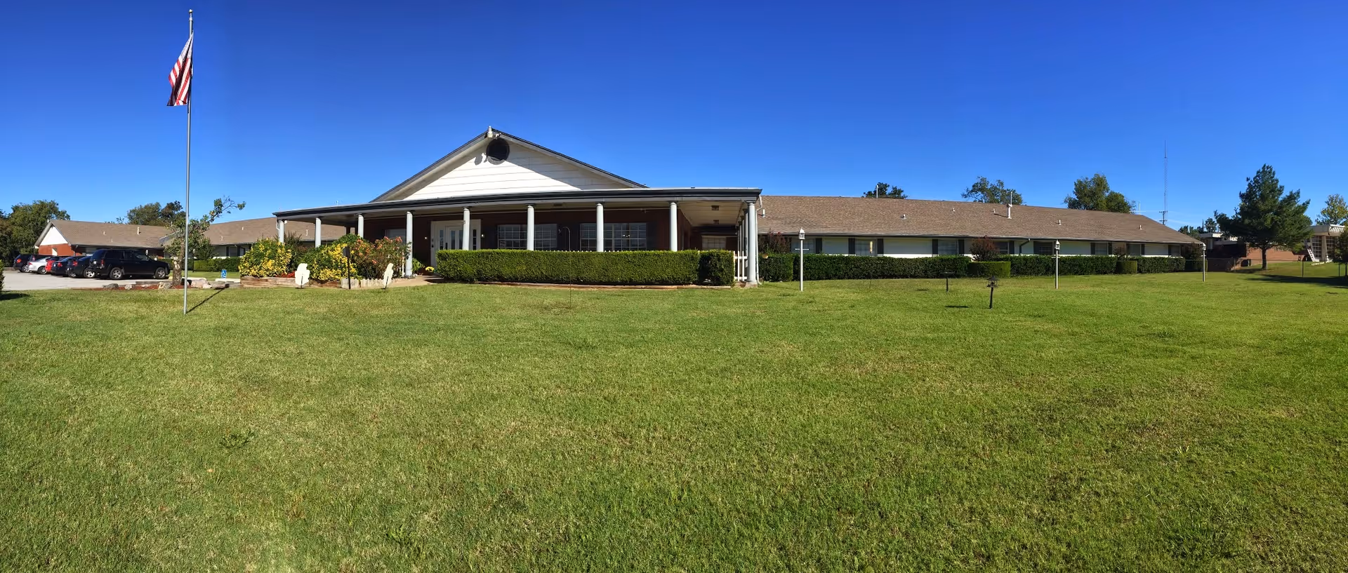 Wide exterior view of Sherwood Manor Nursing Home building with a large green lawn in front, an American flag on a flagpole to the left, and a clear blue sky overhead.