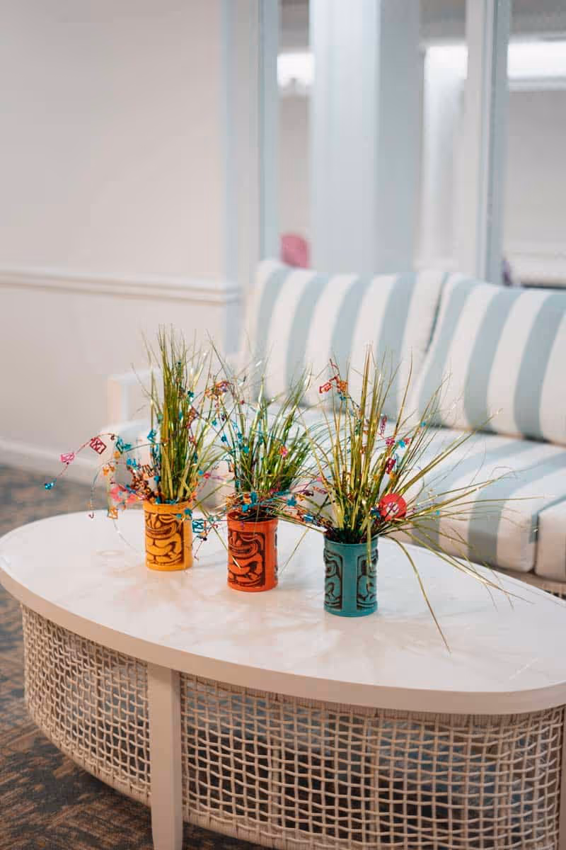 A white oval coffee table with a woven base holds three small colorful pots containing decorative grass and festive wire decorations. In the background, there is a white and light blue striped cushioned sofa against a light-colored wall.