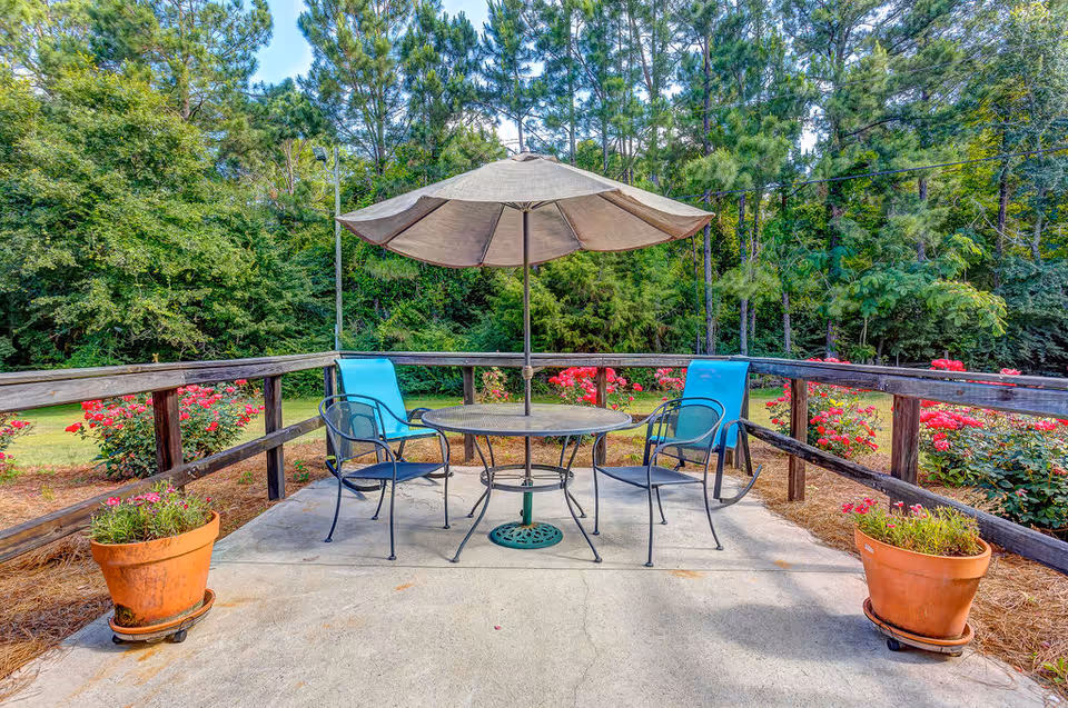 Outdoor patio with a round table, umbrella, four chairs and potted plants overlooking a garden and trees.