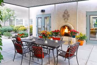 Covered outdoor patio area with a black metal table and six chairs with red cushions. The patio features a built-in fireplace with a fire burning, decorative wall art above it, and potted plants with colorful flowers around the space. Glass doors lead to the interior of the building, and greenery is visible in the background.