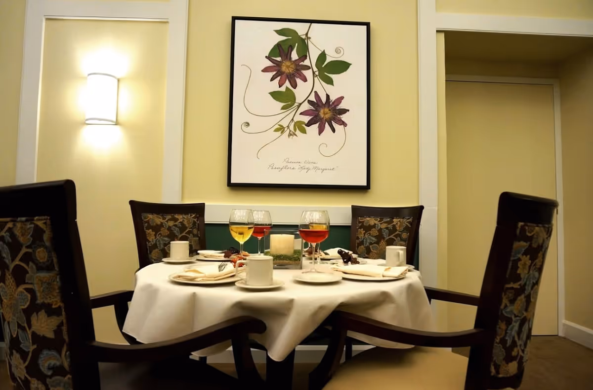 A round dining table set for four with plates, glasses and a centerpiece beneath framed floral artwork in a dining room.