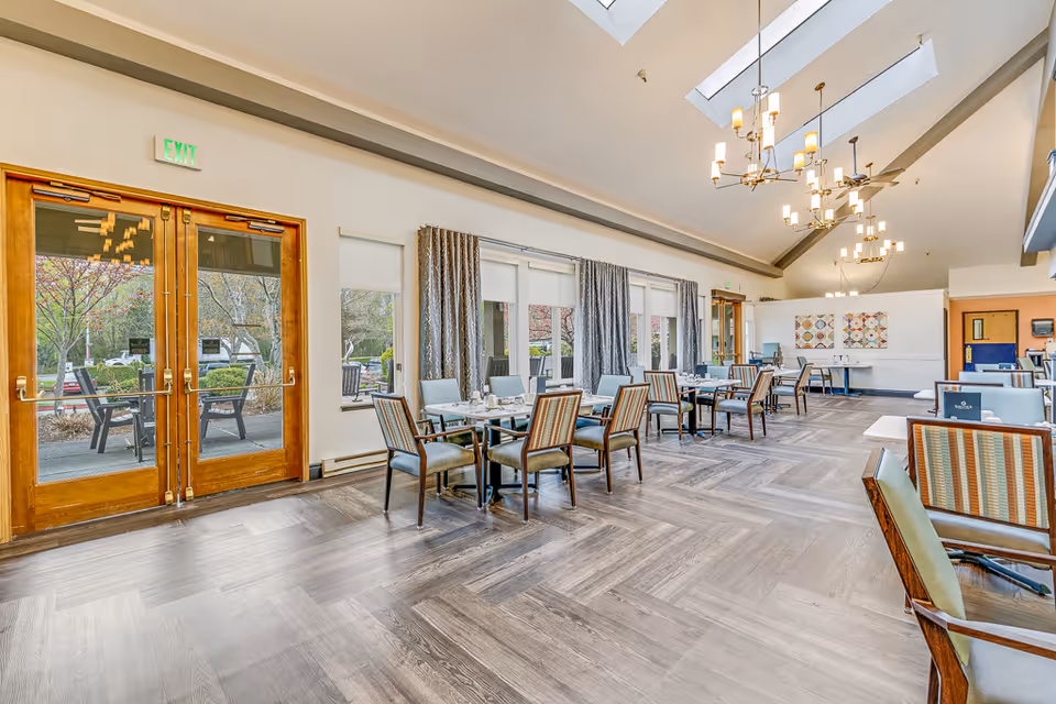 A spacious dining room in Solstice Senior Living at Bellingham featuring multiple tables with chairs arranged neatly. Large windows with curtains allow natural light to enter, and there are double wooden doors leading outside. The room has a high ceiling with modern chandeliers and a patterned wood floor.