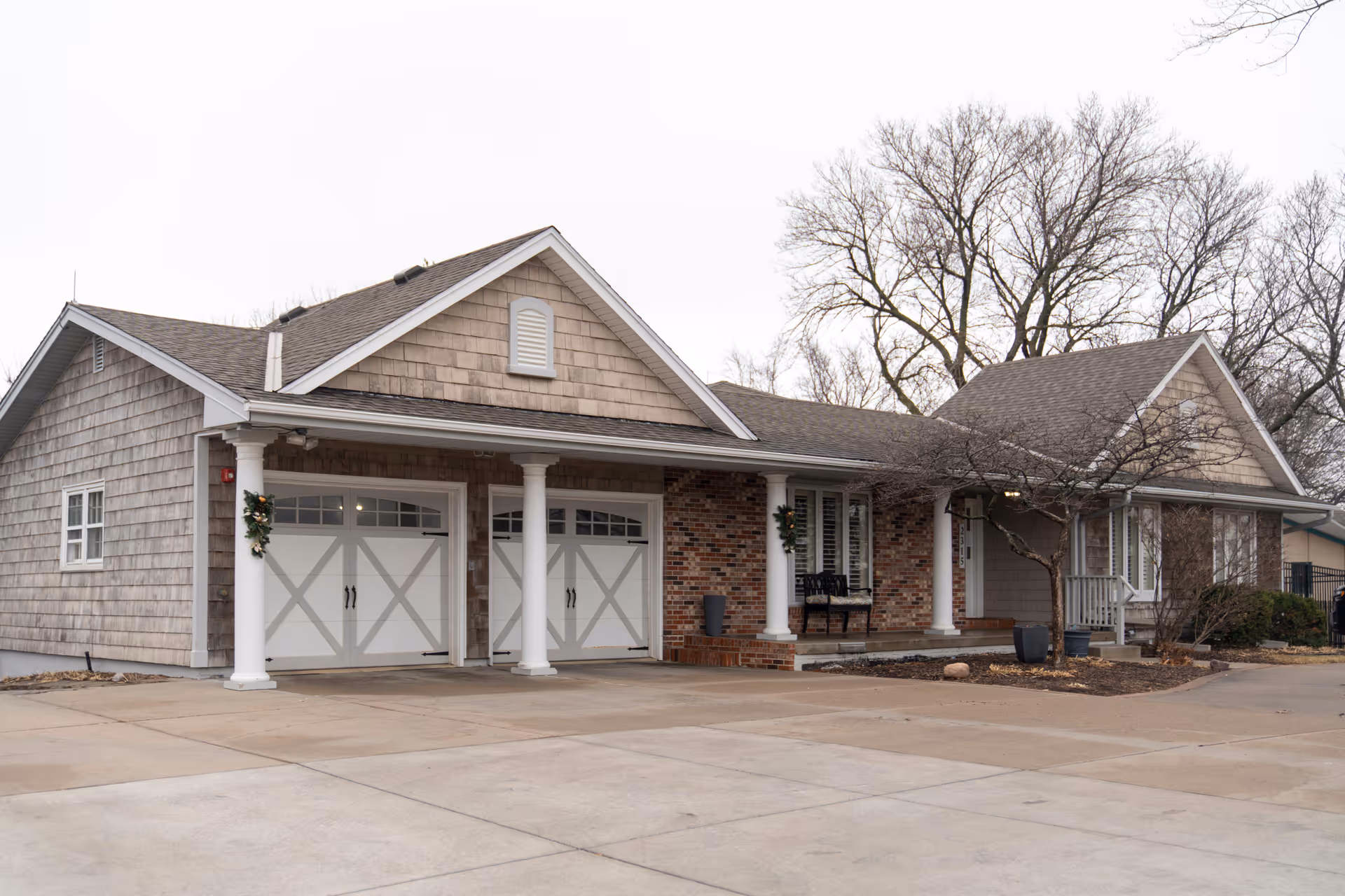 Exterior view of a single-story residential building with a double garage featuring white doors with crossbuck design. The building has beige siding, brick accents, white columns supporting a covered porch, and leafless trees around the property. The driveway and walkway are visible in the foreground.