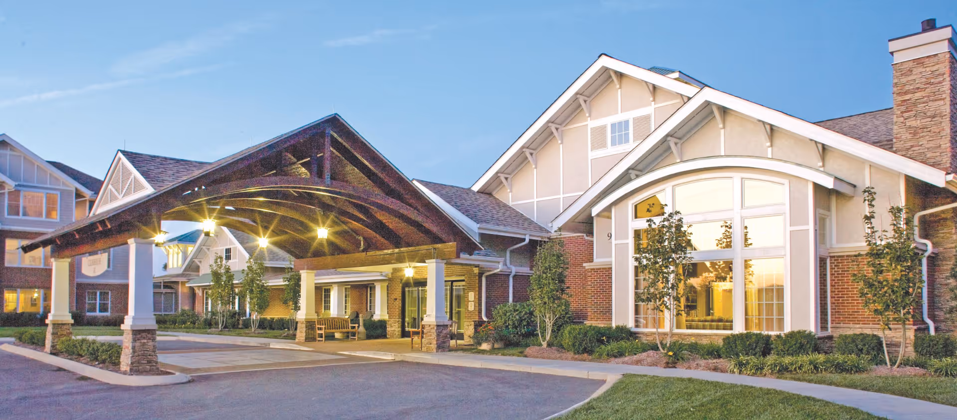 Front entrance of a senior living building with a lit covered porte-cochere, large windows, and landscaped grounds at dusk.