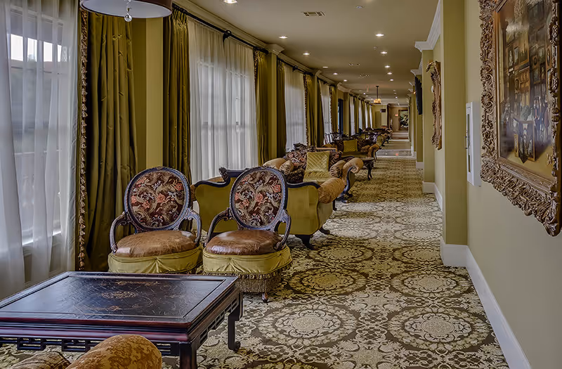 A long, elegant hallway with large windows covered by sheer white curtains and green drapes. The hallway is furnished with ornate chairs and sofas upholstered in patterned fabric, arranged along the wall. The floor is covered with a decorative carpet featuring intricate designs. A framed painting hangs on the right wall.