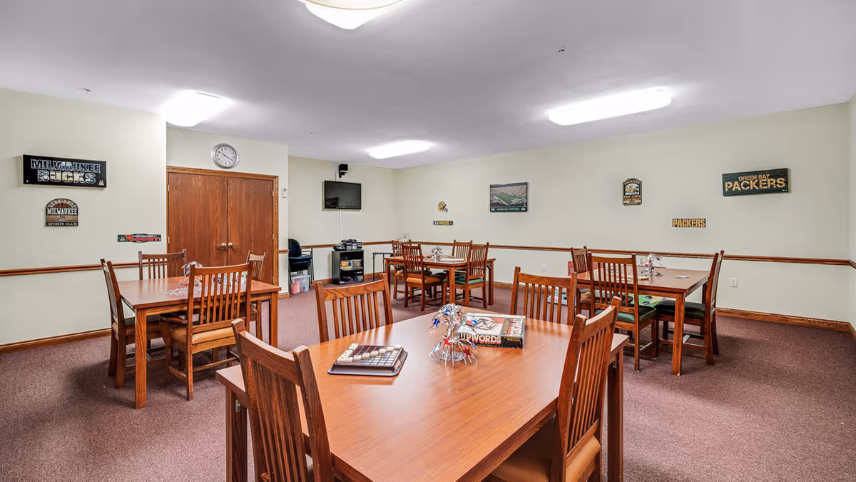 Communal activity/dining room with wooden tables and chairs, a wall-mounted TV, and sports-themed decor on the walls.