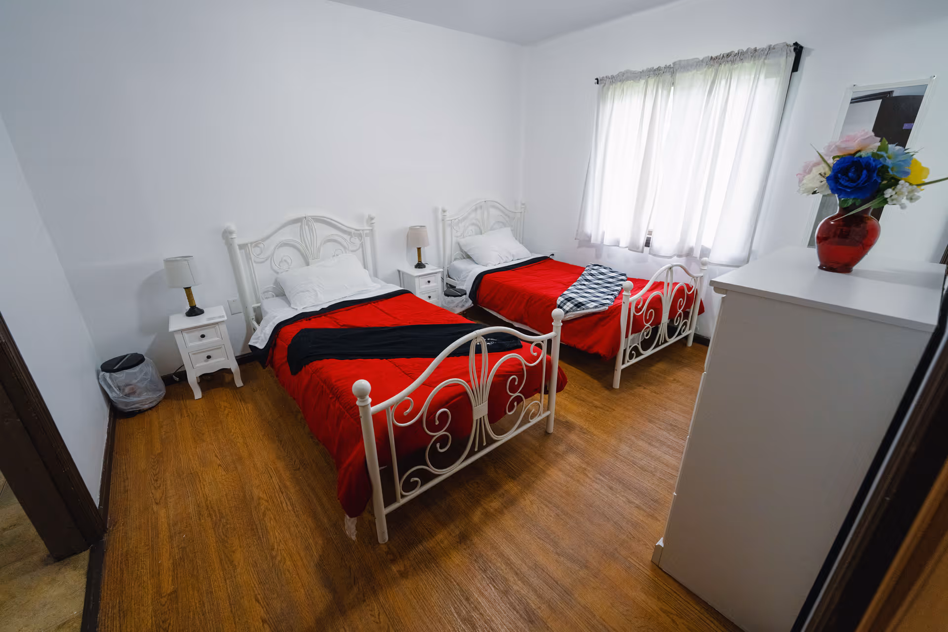 A bright bedroom with two white metal twin beds, each made with red bedspreads and white pillows. Between the beds is a small white nightstand with a lamp. There is a white dresser with a vase of colorful flowers on top near a window with sheer white curtains. The floor is wooden, and the walls are painted white.