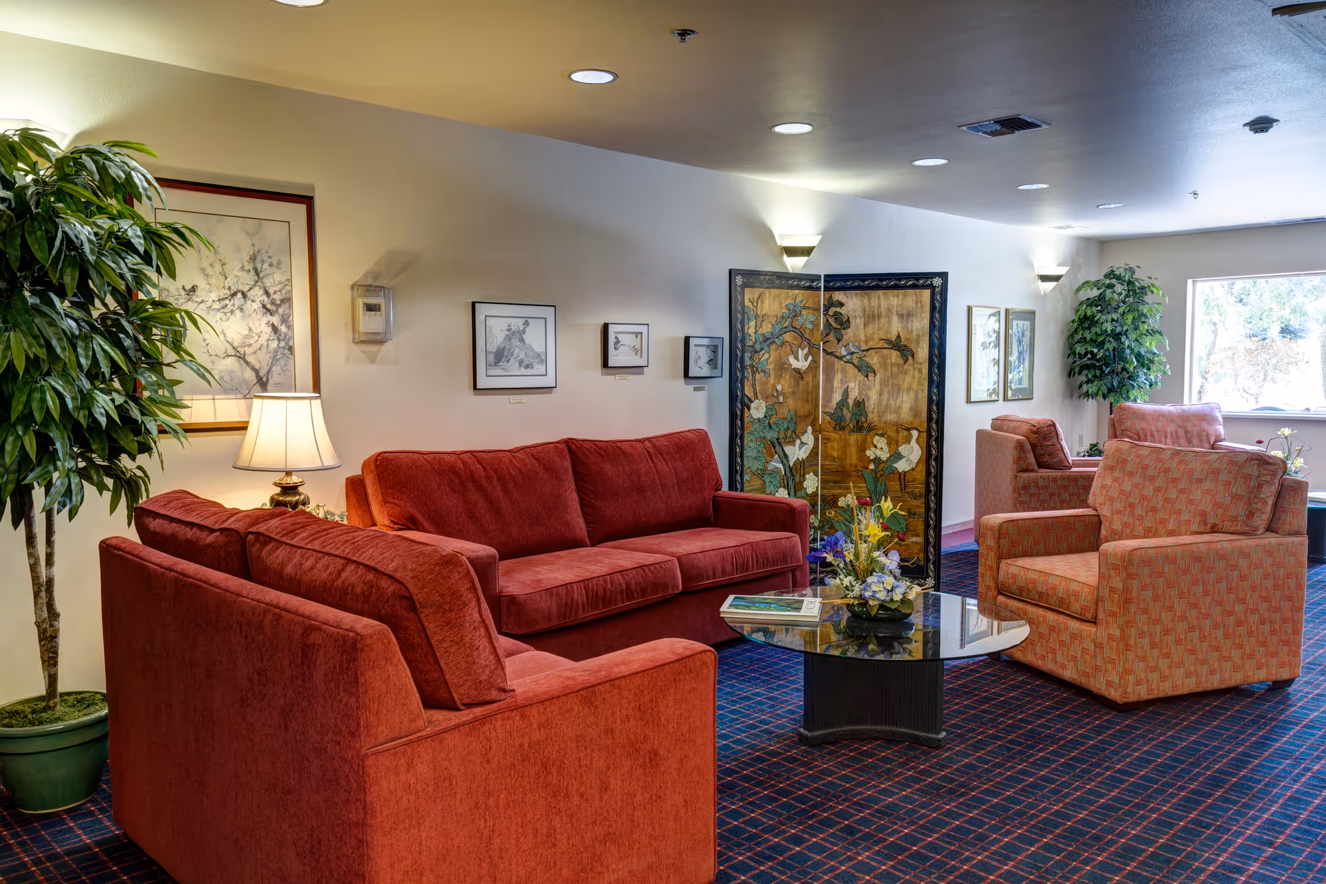A cozy lounge with red sofas and armchairs arranged around a glass coffee table in a well-lit communal room.