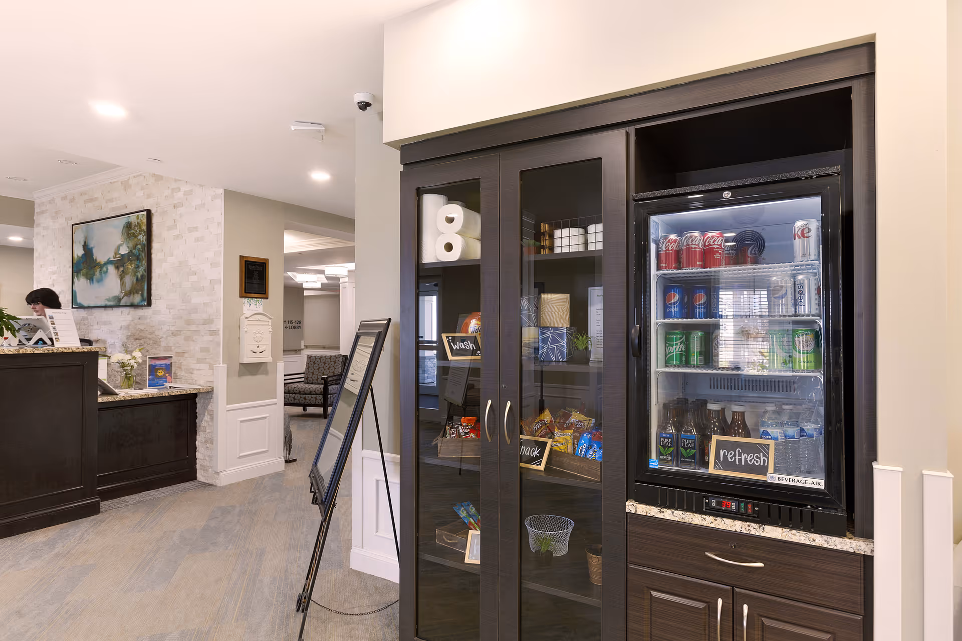 Interior view of a senior living facility reception area with a dark wood snack and beverage cabinet on the right, stocked with snacks and drinks including soda cans and bottled beverages. A reception desk with a person behind it is visible on the left, along with a painting on a light-colored brick wall and seating area in the background.