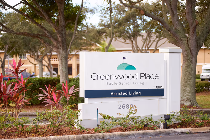 Outdoor view of a white sign for Greenwood Place Eagle Senior Living Assisted Living, surrounded by plants and trees with a building and parked cars in the background.