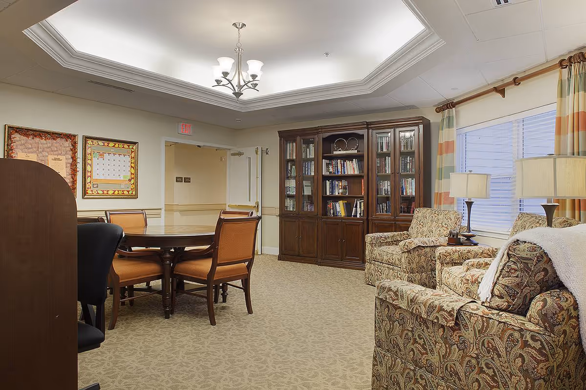 A cozy interior room with a round wooden table surrounded by six chairs, a large wooden bookshelf filled with books, two patterned armchairs, a matching sofa with a white throw blanket, two table lamps on side tables, and a window with striped curtains. The ceiling has a decorative tray design with a chandelier light fixture.
