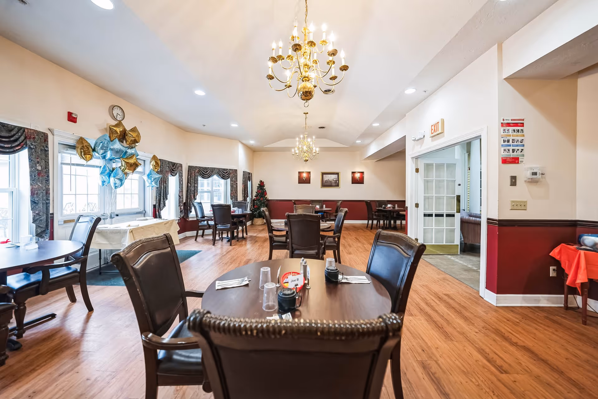 Interior view of the dining room at Seneca Lake Terrace, featuring wooden tables, chairs, and decorative balloons. A Christmas tree is visible in the corner, and large windows allow natural light to fill the space.