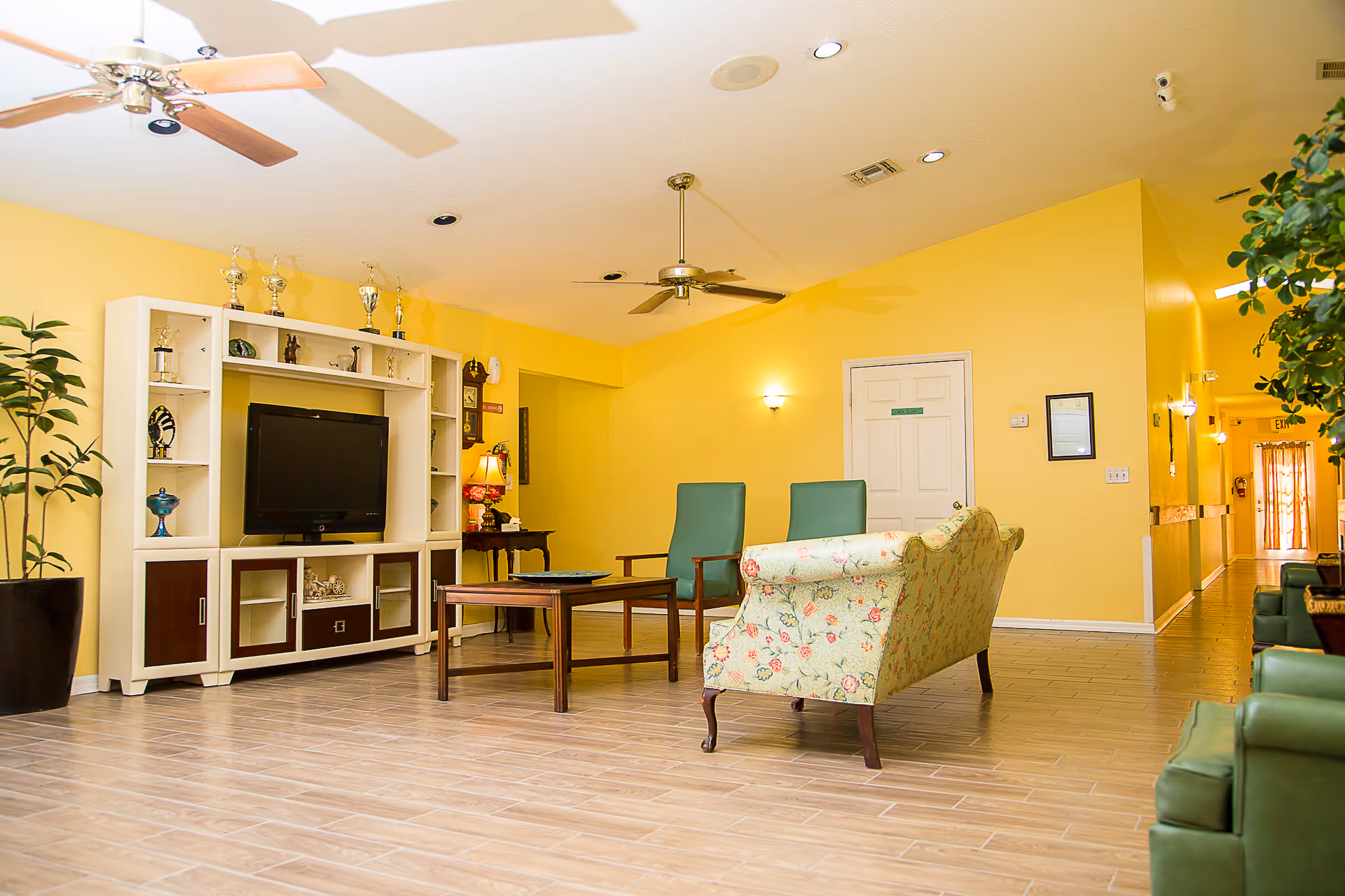 A bright and spacious living room area with yellow walls and light wood flooring. The room features a white entertainment center with a flat-screen TV and several trophies on top. There are two green chairs, a floral patterned sofa, and a wooden coffee table in the center. Ceiling fans and recessed lighting are visible on the ceiling. Potted plants are placed on the sides, and a hallway extends to the right with an exit door at the end.