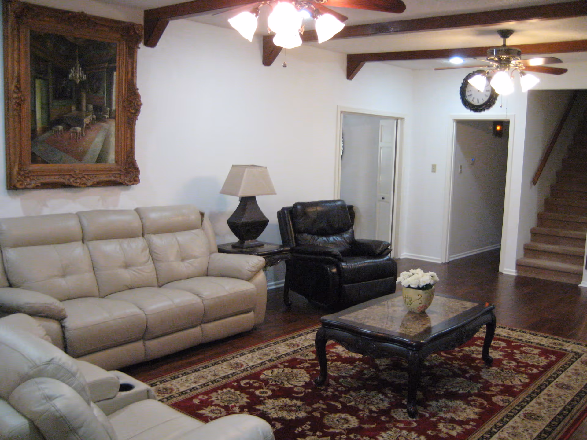 A living room with beige leather sofas, a black leather armchair, a wooden coffee table with a flower pot on top, a patterned red and beige rug, a table lamp on a side table, a large framed painting on the wall, ceiling fans with lights, and a staircase leading upstairs.