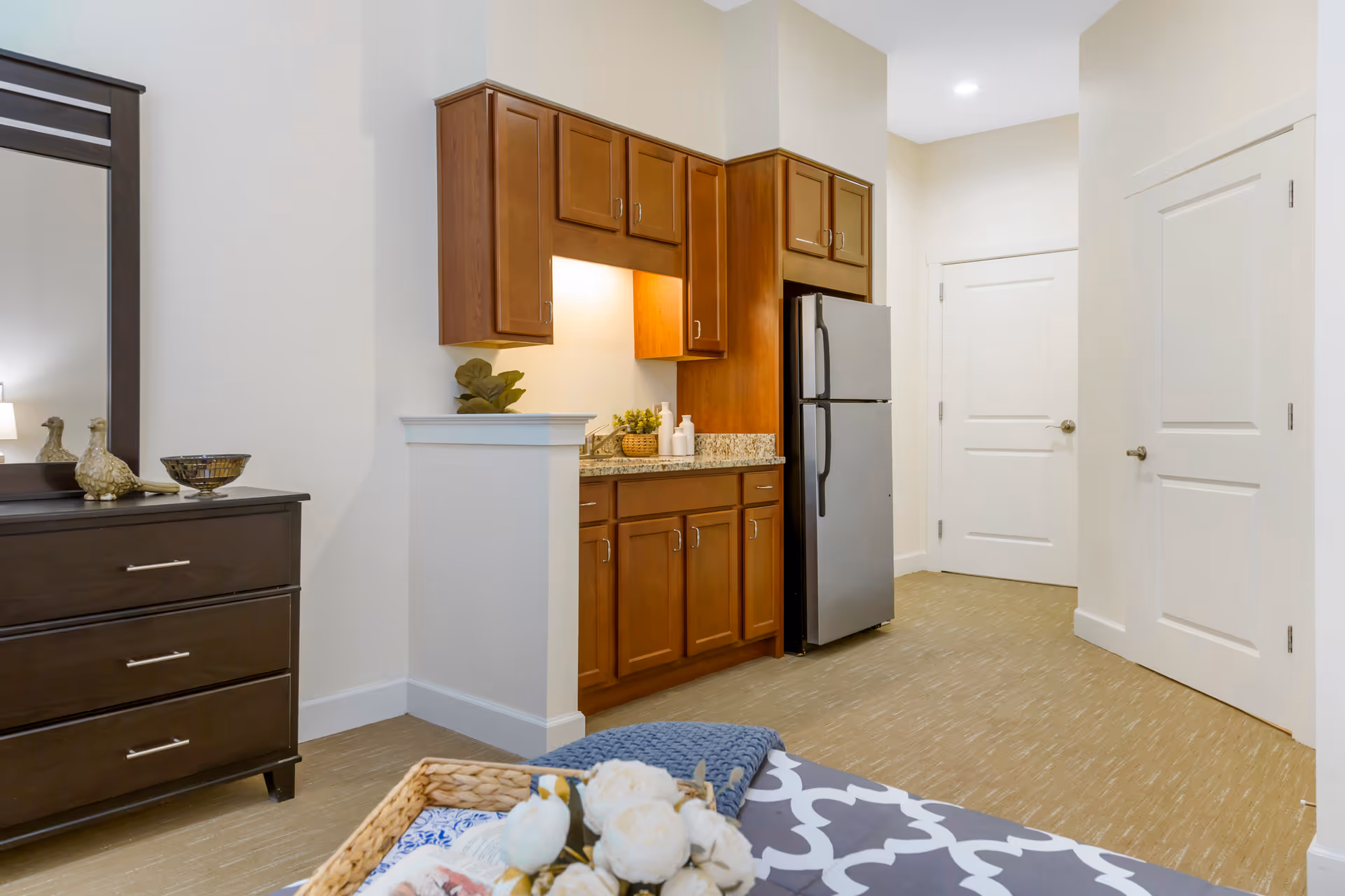 Interior view of a senior living facility room featuring a small kitchenette with wooden cabinets, a granite countertop, and a stainless steel refrigerator. To the left, there is a dark wooden dresser with decorative items on top and a large mirror above it. The foreground shows part of a bed with a patterned bedspread and a tray with flowers and a blanket. The room has light-colored walls and flooring, with two white doors visible in the background.