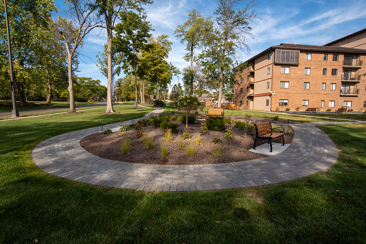 Circular paved garden path with benches and planted landscaping in front of a multi-story brick senior living building under a blue sky.