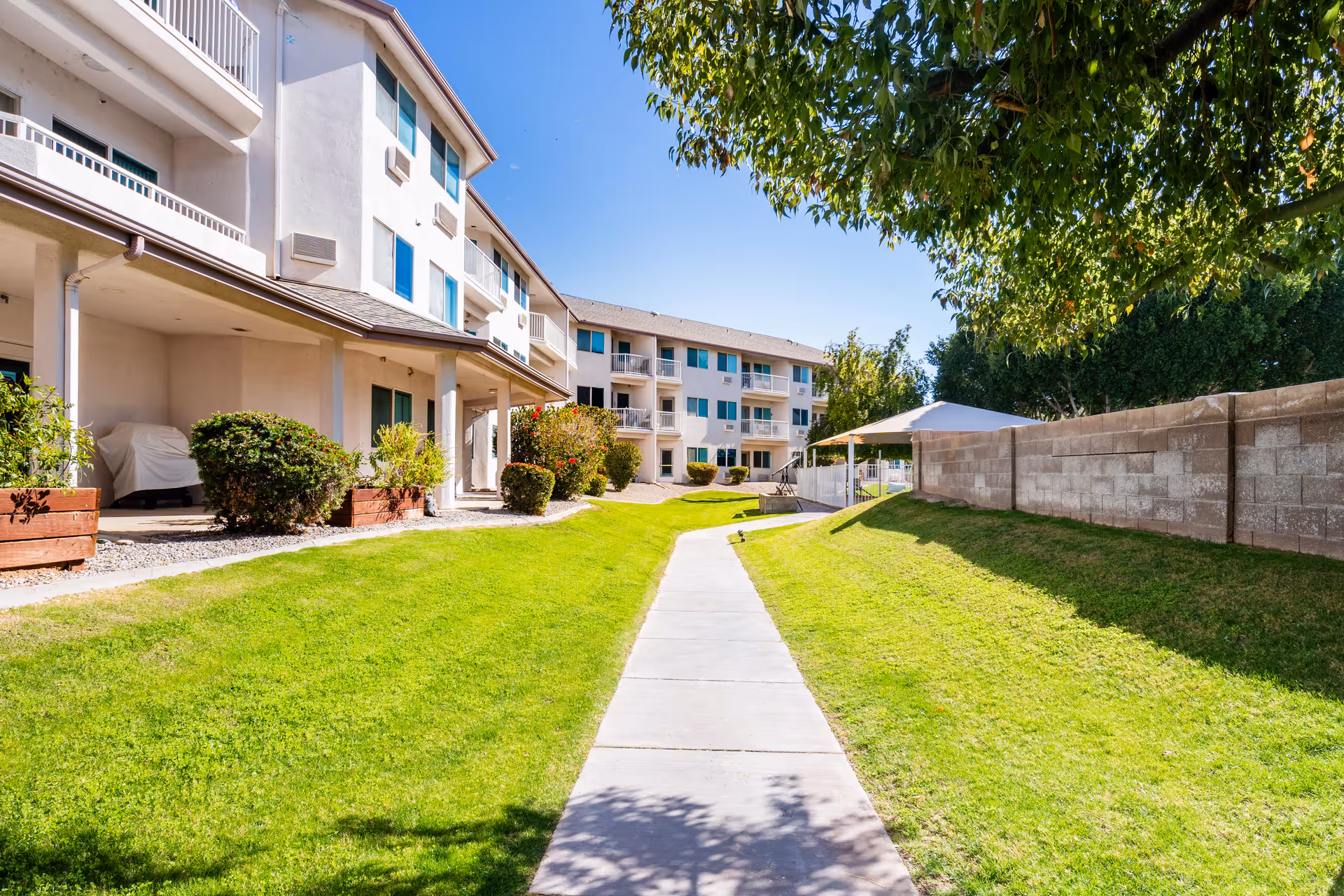 A sunny outdoor pathway lined with green grass and bushes leading to a multi-story residential building with balconies and windows. There is a shaded seating area with a canopy on the right side and trees providing some shade.