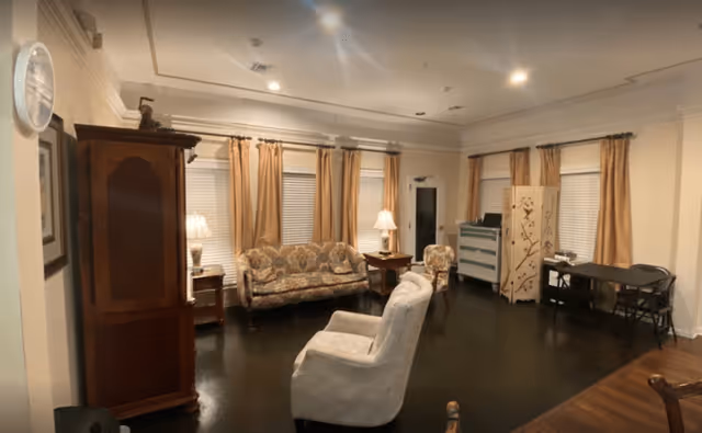 A cozy living room with dark hardwood floors, beige walls, and multiple windows covered with beige curtains. The room features a floral patterned sofa, a white armchair, a wooden armoire, two side tables with lamps, a small desk with a chair, and a decorative folding screen. The ceiling has recessed lighting and crown molding.