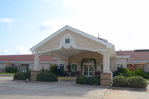Front exterior view of a single-story building with a covered entrance supported by columns, surrounded by bushes and greenery under a partly cloudy sky.