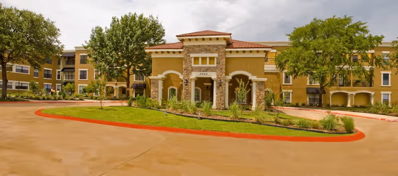 Front exterior view of a three-story assisted living and memory care facility with a central entrance featuring stone accents and a red-tiled roof, surrounded by landscaped greenery and trees under a cloudy sky.