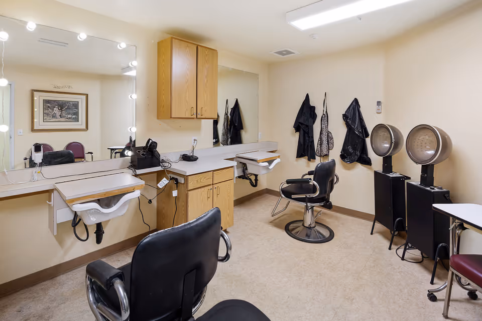 Interior view of a hair salon area in a senior living facility with two black salon chairs, two hair drying stations, a large mirror with lights around it, a small sink, wooden cabinets, and hair styling tools. Aprons hang on the wall and a framed picture is reflected in the mirror.