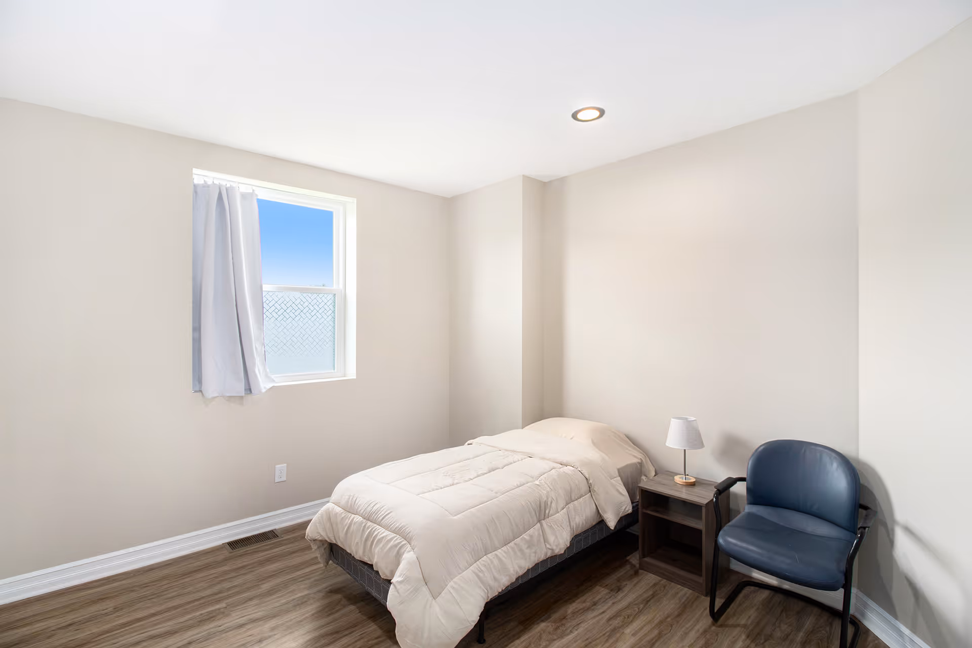 A simple bedroom with a single bed, nightstand and lamp, a blue chair, and a window with a curtain.