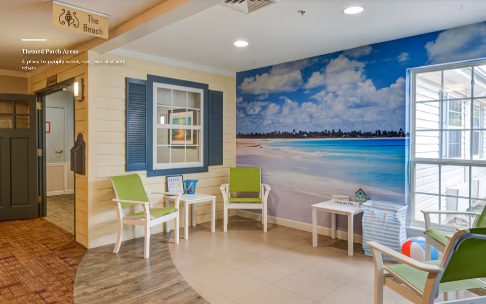 Indoor seating area designed to resemble a beach porch with green cushioned chairs, small white tables, a large wall mural of a beach scene, and a window letting in natural light. The area has a sign labeled 'The Beach' and a cozy, inviting atmosphere.