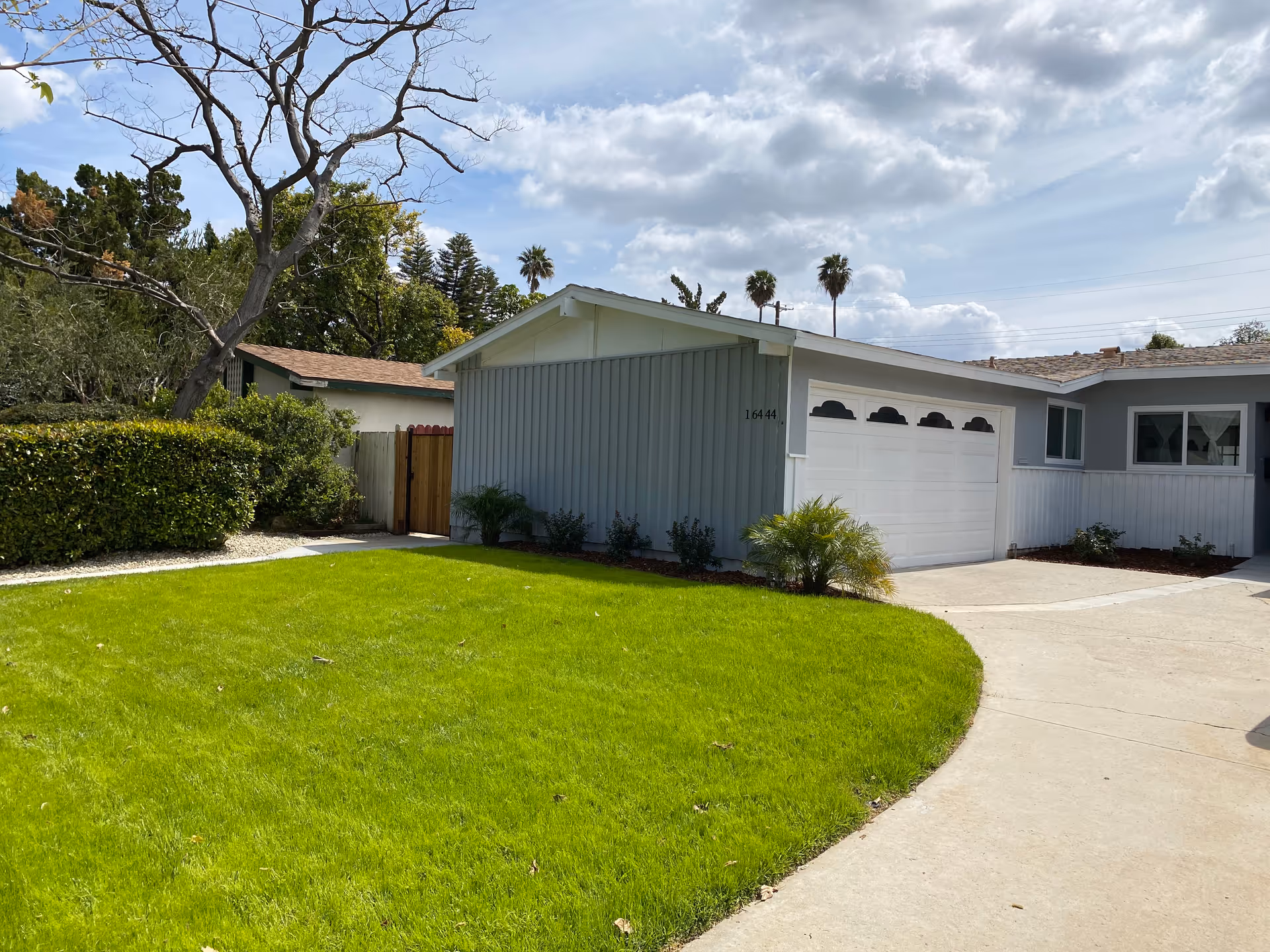 Exterior view of a single-story residential building with a gray and white facade, a white garage door, and a well-maintained green lawn in front. There are some bushes and small plants near the building, a tree with bare branches, and a partly cloudy sky overhead.
