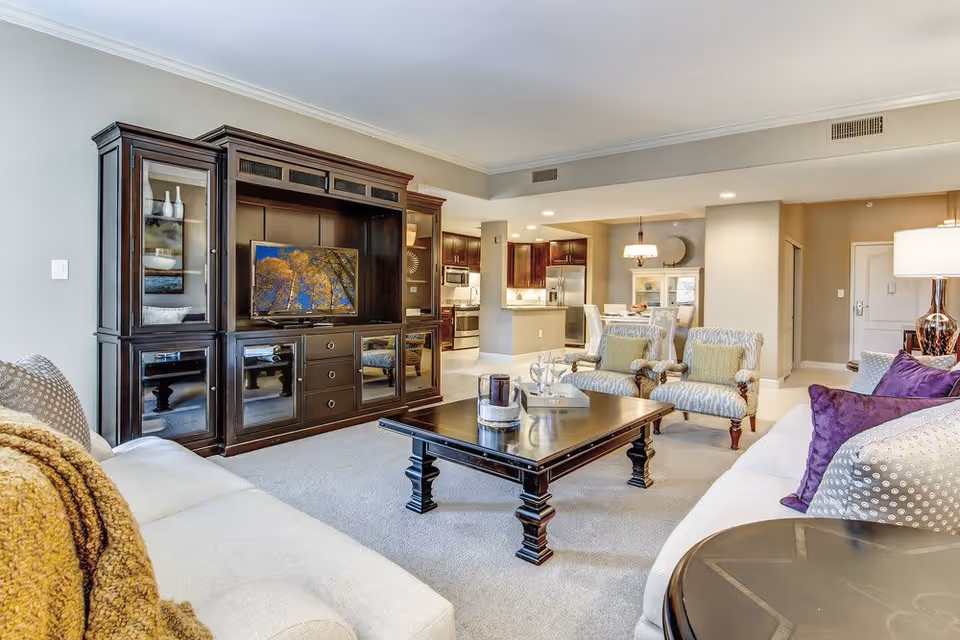 A spacious living room with a large dark wood entertainment center holding a flat-screen TV, two patterned armchairs with green cushions, a dark wood coffee table with decorative items, and two light-colored sofas with purple and patterned pillows. The room opens into a kitchen and dining area with modern appliances and a dining table with chairs.