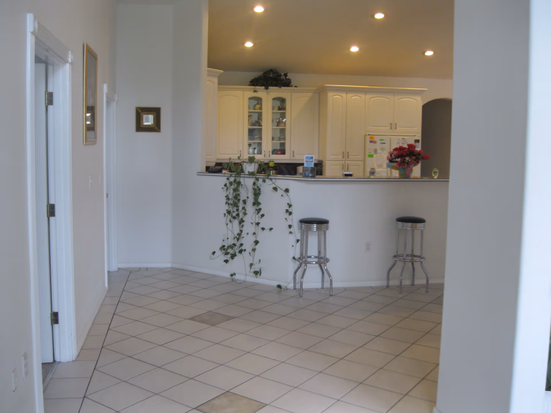 Interior view of a kitchen area with white cabinets, a refrigerator covered with notes, and a counter with two metal bar stools. There is a plant hanging over the counter and a vase with flowers on the countertop. The floor is tiled with a diamond pattern, and the walls are painted white.