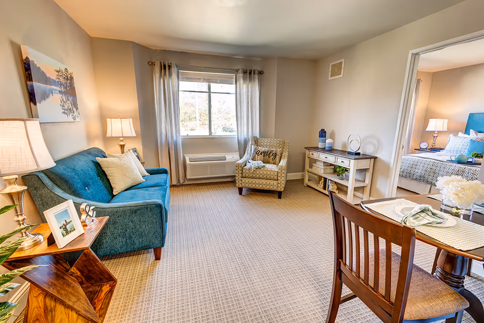 A cozy living room with a teal sofa, a patterned armchair with a 'family' pillow, a wooden side table with framed photos and a lamp, a window with curtains, a wooden console table with decorative items, and a dining table with chairs. A bedroom with a bed and nightstand is visible through an open doorway.