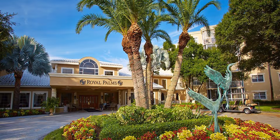 Front exterior view of the Royal Palms senior living facility with a landscaped garden featuring palm trees, colorful flowers, and a bronze sculpture of two cranes. The building has a beige facade with white roof tiles and large windows under a clear blue sky.