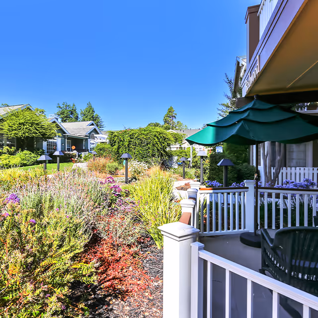 Sunny landscaped courtyard with a porch, green patio umbrella, and neighboring residential buildings.