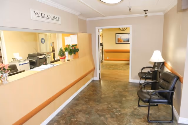 Reception area of a senior living facility with a welcome sign above a check-in window and chairs with a lamp along the hallway.