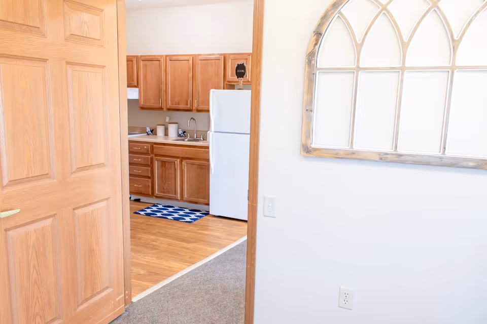 View through an open wooden door into a kitchen area with wooden cabinets, a white refrigerator, a sink, and a blue patterned rug on a wooden floor. On the right wall, there is a decorative window frame with an arched design.