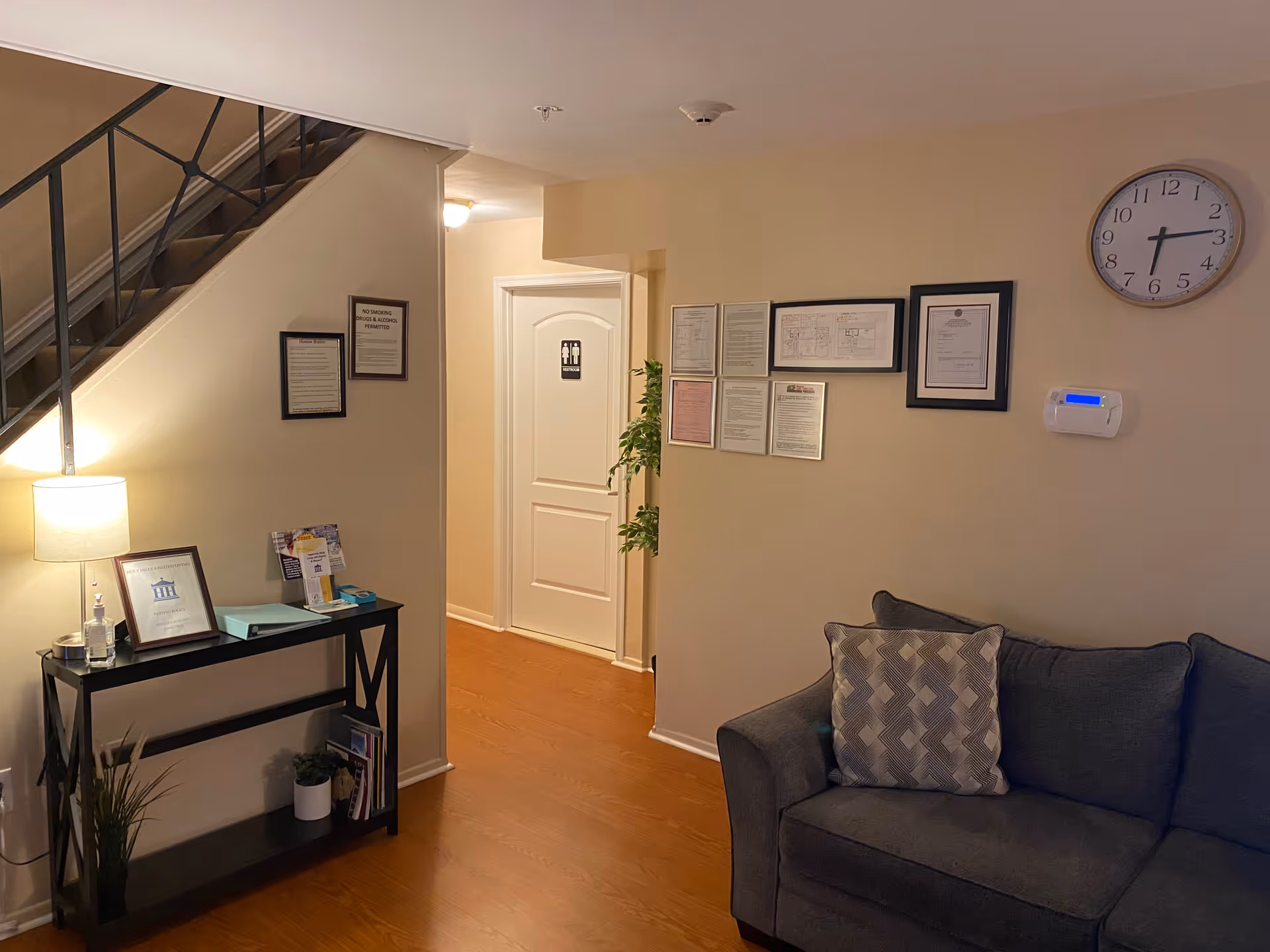 Cozy lobby area at Holy Hills Assisted Living with couch, clock, framed certificates, and stairs.